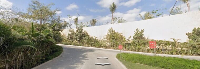 Street view of the primary entrance road to LUDŌ at VidantaWorld, featuring the massive white security perimeter wall and the first left-hand turn toward the BON Park Station 2 Uber drop-off.